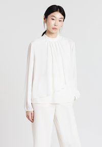 Weiße Bluse mit langen Ärmeln, gerüschtem Ausschnitt und drapierter Vorderseite. Glattes, leichtes Material mit einem subtilen Glanz.