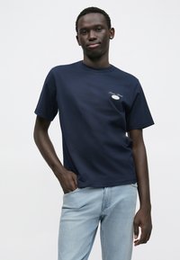 Μπλε ναυτικό βαμβακερό t-shirt με κοντά μανίκια, στρογγυλή λαιμόκοψη και μικρό κεντημένο λογότυπο στην αριστερή πλευρά. Φορεμένο με ανοιχτόχρωμα μπλε τζιν.