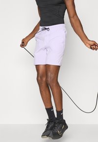 Shorts de sport violets clairs avec une taille élastique, un cordon de serrage noir, des poches latérales et un petit logo. Portés avec un débardeur noir et des chaussures.