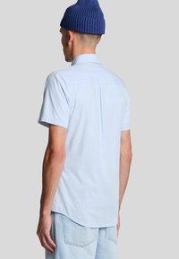 Camicia a maniche corte con bottoni di colore azzurro chiaro, caratterizzata da un sottile motivo a quadri. Il tessuto è traspirante e presenta un colletto classico e un dettaglio con piega nella parte posteriore.
