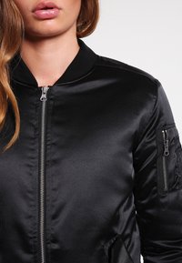Veste bombardier noire en tissu satin, avec fermeture éclair, col côtelé et poche latérale. Texture lisse avec une finition brillante.