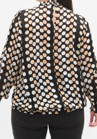 Blouse noire transparente avec un grand motif hexagonal en tons blanc et beige. Dotée de manches longues, d'un col haut et d'un ourlet froncé.
