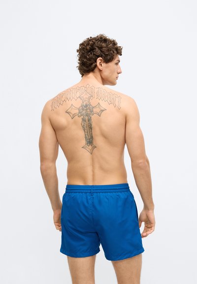 Siniset uimashortsit, jotka on valmistettu sileästä, kevyestä materiaalista ja joissa on joustava vyötärönauha. Takana on yksityiskohtainen tatuointikuvio.