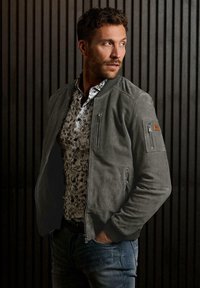 Grau Wildleder Bomberjacke mit Reißverschlusstaschen, gerippten Bündchen und einem designten Akzent am Ärmel. Tragen über einem floral gemusterten Hemd.