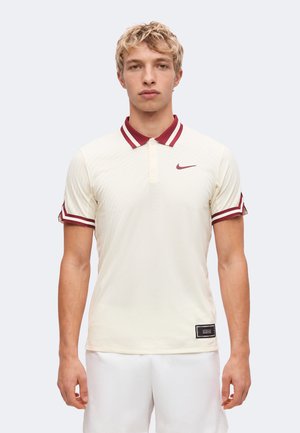 Jeune homme aux cheveux blonds courts portant un polo crème Nike avec un col et des bandes bordeaux sur les manches, debout devant un fond clair.