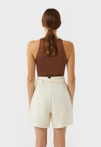 Top marrón sin mangas con un cuello alto, combinado con pantalones cortos crema de talle alto que presentan un cinturón y una tela texturizada.