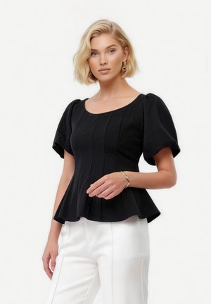 Blonde Frau mit goldenen Creolen, einem enganliegenden schwarzen Oberteil mit Puffärmeln und Schößchen sowie weißen, hoch taillierten Hosen vor einem weißen Hintergrund.