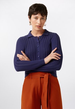 Marineblauer Strickpullover mit Polo-Kragen, Rippmuster und Knopfleiste, kombiniert mit orangefarbenen, hochtaillierten Hosen mit einem gebundenen Gürtel.
