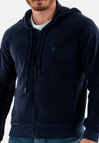 Marineblauwe fleece hoodie met rits, voorzien van een trekkoordkap, raglanmouwen en een klein logopatch op de borst. Heeft ribgebreide manchetten en zoom.