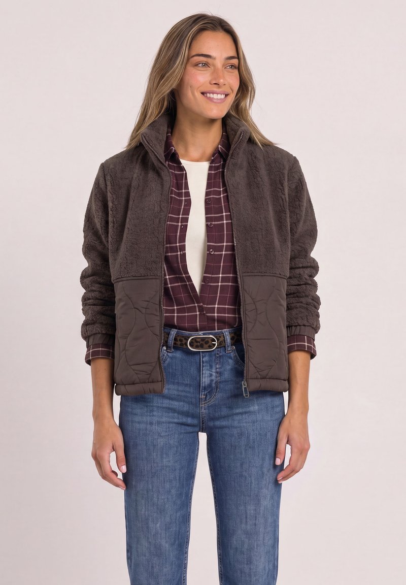 Woman wearing a brown fleece jacket over a burgundy plaid shirt and blue jeans, smiling and standing against a light background.