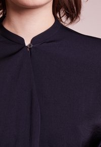 Tummansininen kauluspaita, jossa on napitus kaula-aukon kohdalla; kevyt kangas, jossa on sileä pinta ja minimalistinen muotoilu.