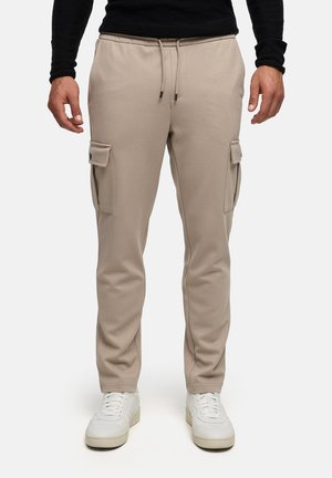 Pantalones cargo beige con bolsillos laterales con solapa, cintura elástica con cordones, usados con zapatillas blancas y una camiseta negra de manga larga.