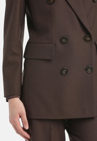 Blazer marrón de doble botonadura con tejido texturizado, que presenta grandes botones oscuros y un bolsillo lateral. Puños en un tono contrastante visibles.