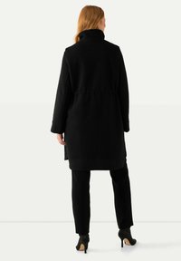 Cappotto nero, oversize, con collare alto e vita con cintura, realizzato in materiale testurizzato. Presenta maniche lunghe e scende sotto il ginocchio.