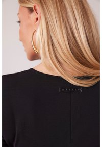 Czarna bluzka o gładkiej fakturze, z okrągłym dekoltem, subtelnymi detalami stikacjami oraz metką z logo z tyłu. Widoczne złote kolczyki w kształcie kół.
