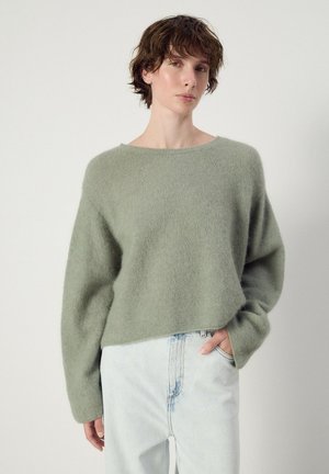 Person mit kurzen braunen Haaren, die einen lockeren hellgrünen flauschigen Pullover und hellblaue hoch taillierte Jeans trägt, eine Hand in der Tasche.