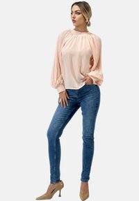 Blusa rosa claro con mangas largas plisadas, combinada con jeans ajustados azules y tacones altos beige, posando de manera relajada.