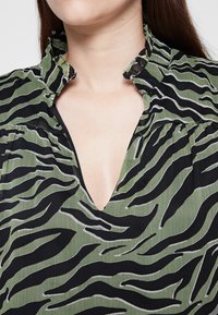 Blusa a righe verdi e nere con scollatura a V profonda e colletto arricciato, realizzata in un tessuto testurizzato e con dettagli arricciati lungo le spalle.