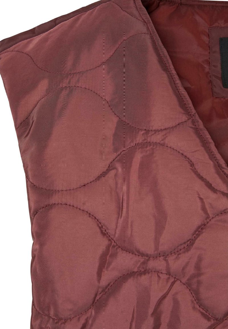 Gilet matelassé dans un bordeaux profond, avec une finition satin lisse et des motifs ondulés arrondis. L'intérieur est d'une teinte unie plus foncée.