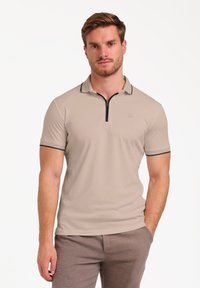Polo de manga corta beige con cierre en la parte delantera, ribete negro en el cuello y un discreto parche de logo, combinado con pantalones marrones texturizados.