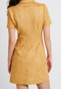 Abito a maniche corte di colore giallo senape realizzato in un morbido tessuto simile alla pelle scamosciata; design aderente con colletto e una lieve texture su tutta la superficie.