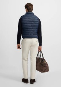 Hombre con chaleco de plumas azul marino, camiseta de manga larga negra y pantalones blancos, sosteniendo un bolso de cuero marrón en la mano derecha, de pie frente a un fondo liso.