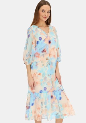 Kobieta ubrana w pastelową, kwiatową sukienkę midi z rękawami do połowy przedramienia zakończonymi bufkami i falbanistym dołem, stojąca na jednolitym tle.