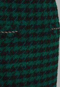 Černozelená látka s vzorem houndstooth, má texturovaný povrch. Obsahuje malé stříbrné akcenty blízko švu.