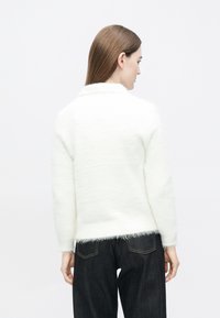 Biely nadýchaný sveter s textúrovaným povrchom, vybavený golierom a rebrovanými manžetami. Kombinovaný s tmavými džínsami s vreckami vzadu.