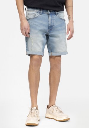 Hellblaue Denim-Shorts mit umgeschlagenen Säumen, die abgenutzte Stellen und ein klassisches Fünf-Taschen-Design aufweisen. Getragen mit beigen Sneakers.