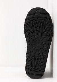 Semelle de chaussure en caoutchouc noir présentant un motif de bande de roulement en zigzag et un logo circulaire au centre. Surface texturée pour l'adhérence et la durabilité.