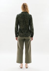 Blusa a maniche lunghe di un verde scuro con motivo leopardato, abbinata a pantaloni ampi color oliva e sandali rosa con cinturini.