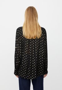 Schwarze durchsichtige Bluse mit goldenen ovalen Punkten, langen Ärmeln, lockerer Passform und einem leicht gerafften Rückendetail. Getragen über blauen Denim-Hosen.