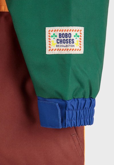 Veste de ski colorée avec une partie supérieure verte, une partie inférieure marron et des poignets élastiques bleus. Présente un logo en patch blanc avec un texte coloré.