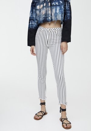 Persona che indossa pantaloni bianchi attillati con righe verticali nere, orli sfrangiati, sandali neri con cinturini e una giacca di jeans cropped tie-dye.