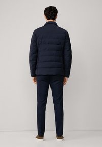 Marineblauwe pufferjack met een gequilt ontwerp, zichtbare stiksels en een kraag, gecombineerd met donkergestreepte broeken en bruine sneakers.