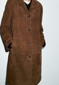 Bruinen suède jas met een knoopsluiting aan de voorkant, een getailleerde kraag en twee voorkruiszakken. Soepele textuur en een getailleerde pasvorm voor een gestructureerd silhouet.