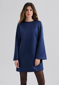 Robe bleu marine avec une texture lisse, des manches cloche longues et un col rond. L'ourlet se situe au-dessus du genou, associée à des collants noirs transparents.