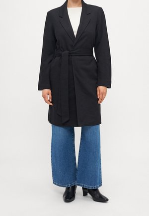 Personne portant un long manteau noir ceinturé, un jean bleu à jambes larges et des bottines noires à talons, debout devant un fond blanc uni.