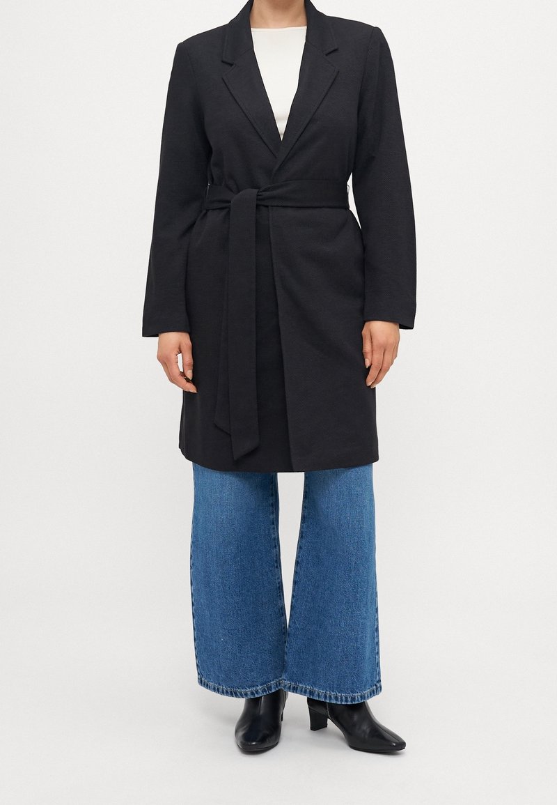 Osoba ubrana w czarny długi płaszcz z paskiem, szerokie niebieskie jeansy i czarne botki na obcasie, stojąca na tle jednolitej białej ściany.