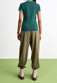 Osoba stojící na zelené podlaze, oblečená v zelené košili s krátkým rukávem, volných olivových kalhotách a béžových kotníkových botách s černými podrážkami.