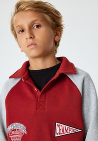 Sudadera roja y gris con cuello, que presenta parches bordados de "Academia de Baloncesto" y "CAMPEÓN". Tejido suave y texturizado con un diseño deportivo.