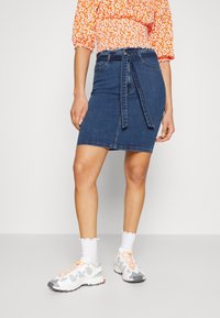 Denim pencil skirt with a blue wash, featuring a tied waist, front pockets, and a knee-length hem. Paired with white sneakers and socks.