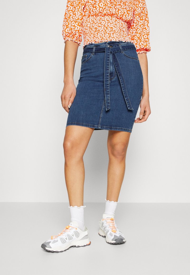 Denim pencil skirt with a blue wash, featuring a tied waist, front pockets, and a knee-length hem. Paired with white sneakers and socks.