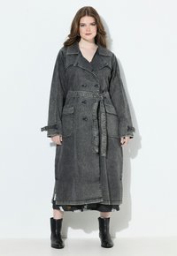 Žena s dlouhými vlasy má na sobě tmavě šedý trench coat s páskem a velkými kapsami a černé kotníkové boty, stojí na bílém pozadí.