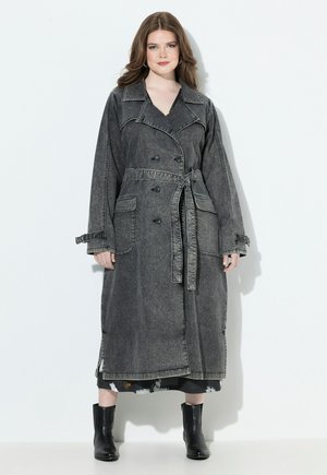 Een vrouw met lang haar draagt een donkergrijze trenchcoat met riem en grote zakken, en zwarte enkellaarzen, terwijl ze tegen een witte achtergrond staat.