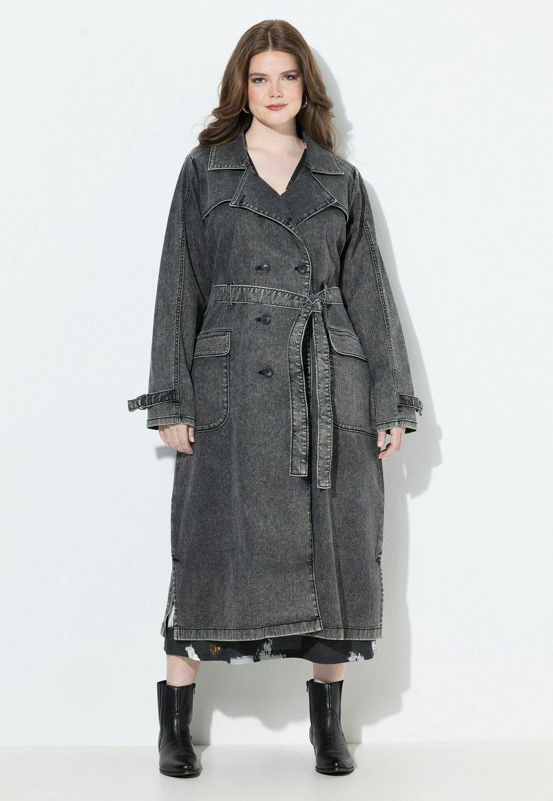 Kobieta z długimi włosami ubrana w ciemnoszary płaszcz trench z szerokimi kieszeniami i czarnymi botkami za kostkę, stojąca na białym tle.