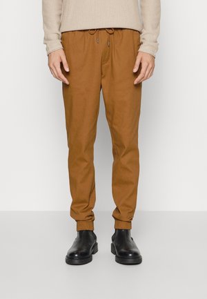 Mann trägt braune Jogginghose mit Kordelzug in der Taille und schwarze Leder-Stiefeletten, steht vor einem schlichten weißen Hintergrund.