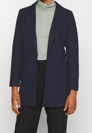 Persoon die een donkerblauw colbert draagt over een lichtgroene geribbelde coltrui en een zwarte broek, tegen een effen lichte achtergrond.