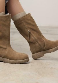 Botas altas de ante marrón con suela texturizada, cremallera lateral y puño de punto en la parte superior, exhibidas sobre una superficie de concreto.